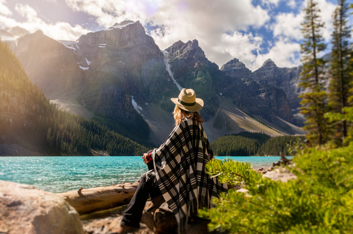 Person in a hat and plaid blanket sitting by a turquoise lake, with towering mountains in the background.