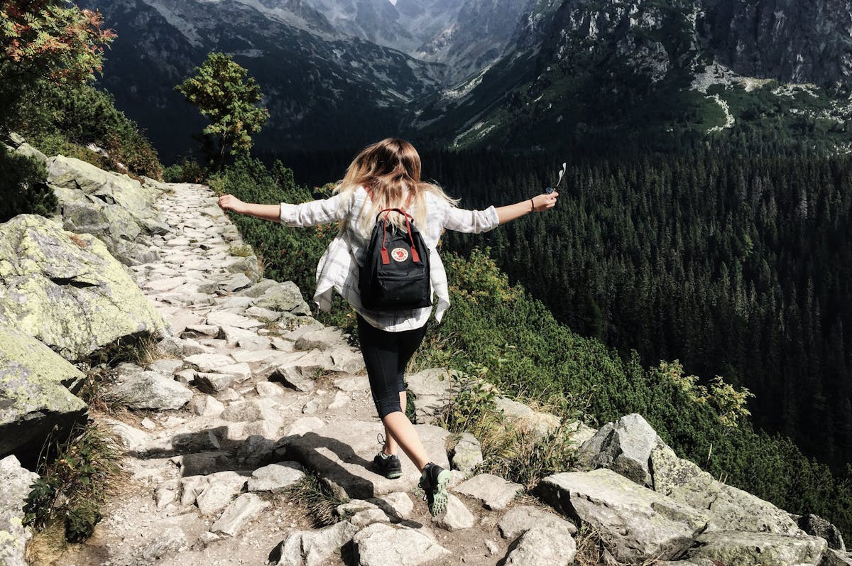 Person hiking down a rocky mountain path, surrounded by dense forest and distant peaks.
