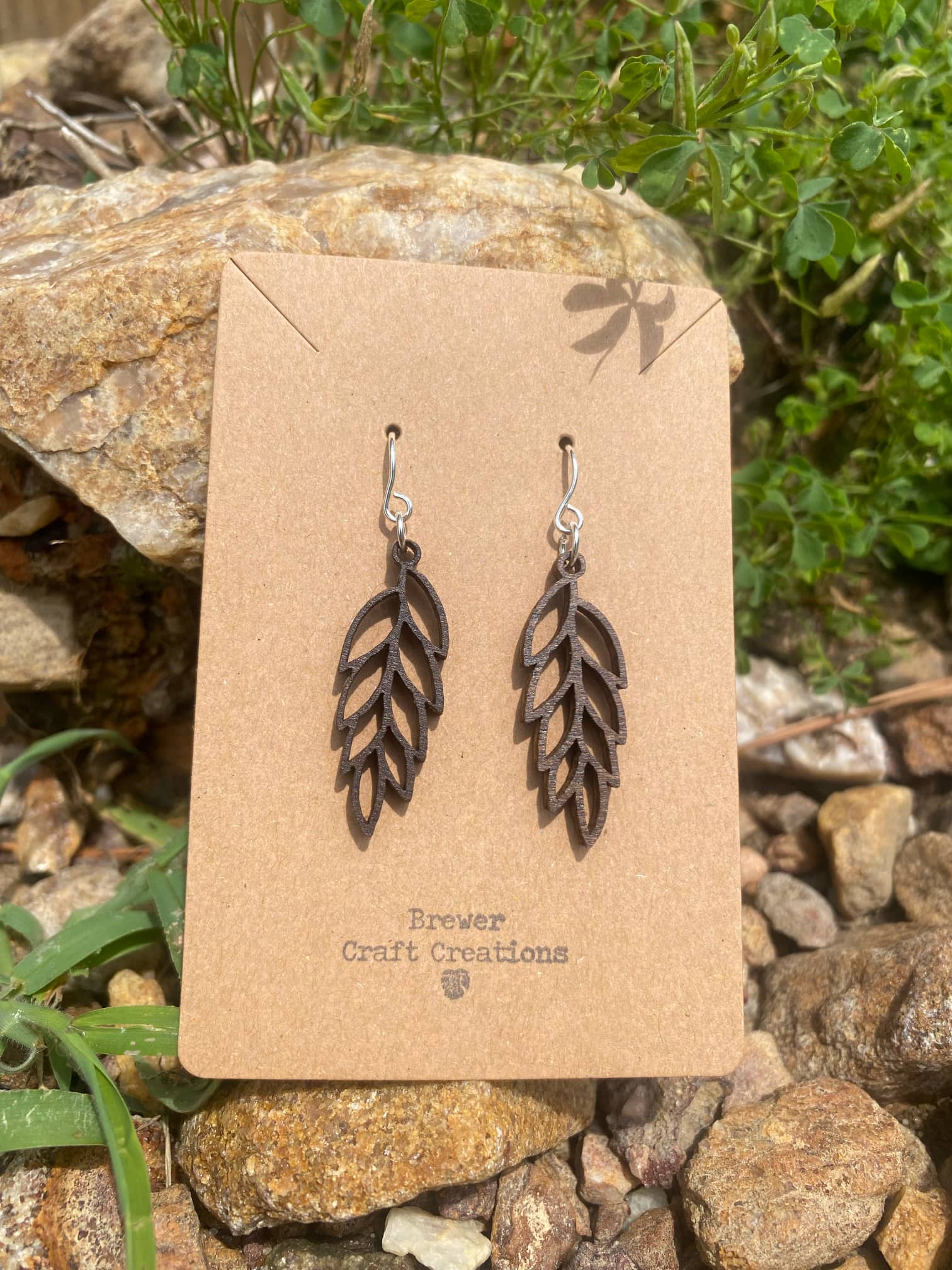 Classic long leaf earring on a cardboard backing being displayed on a rock in front of grass. 