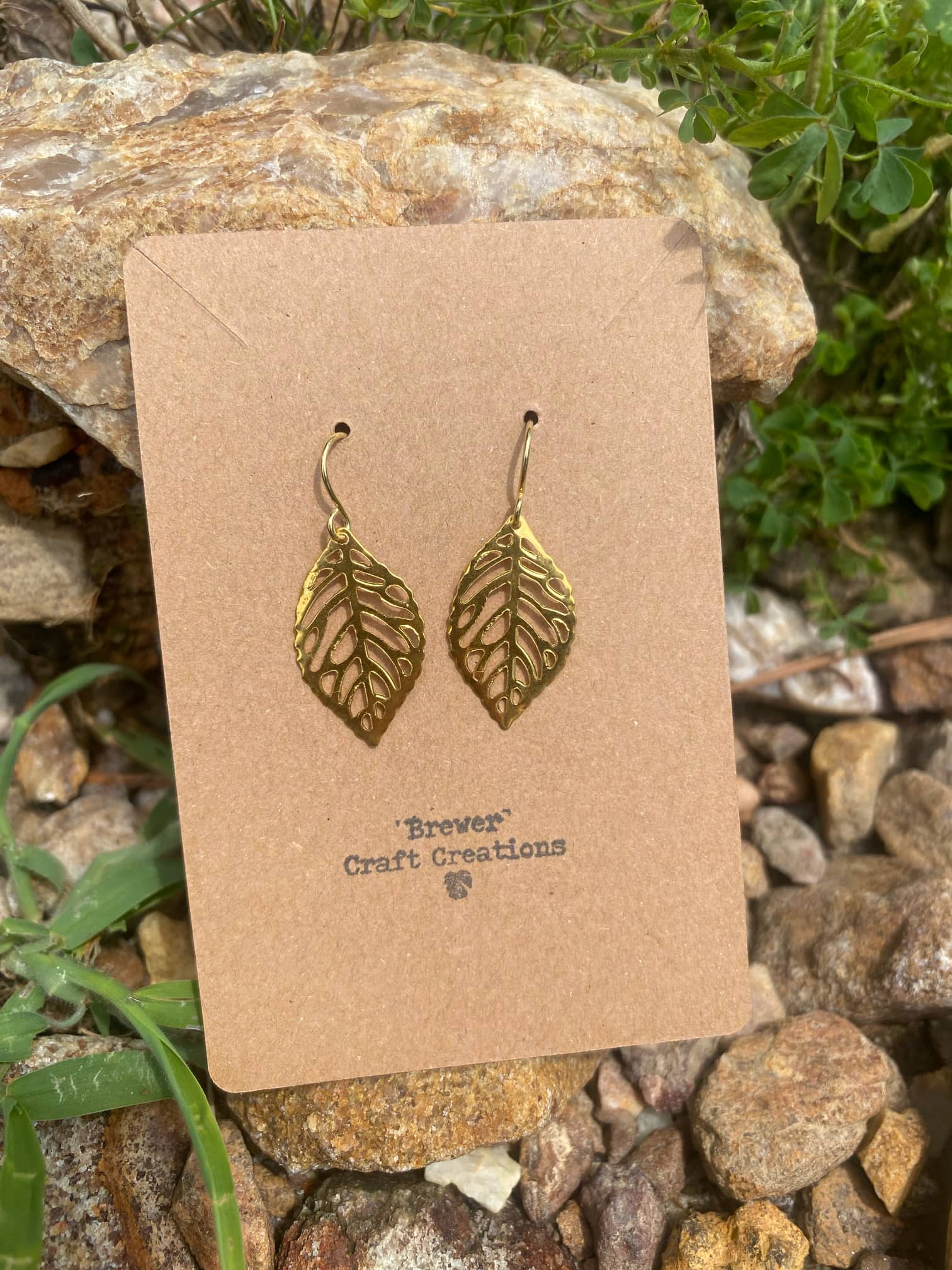 Sol Leaf Foil Earrings on a cardboard backing, gold leaf-shaped dangles, leaning against a rock with a light grass background