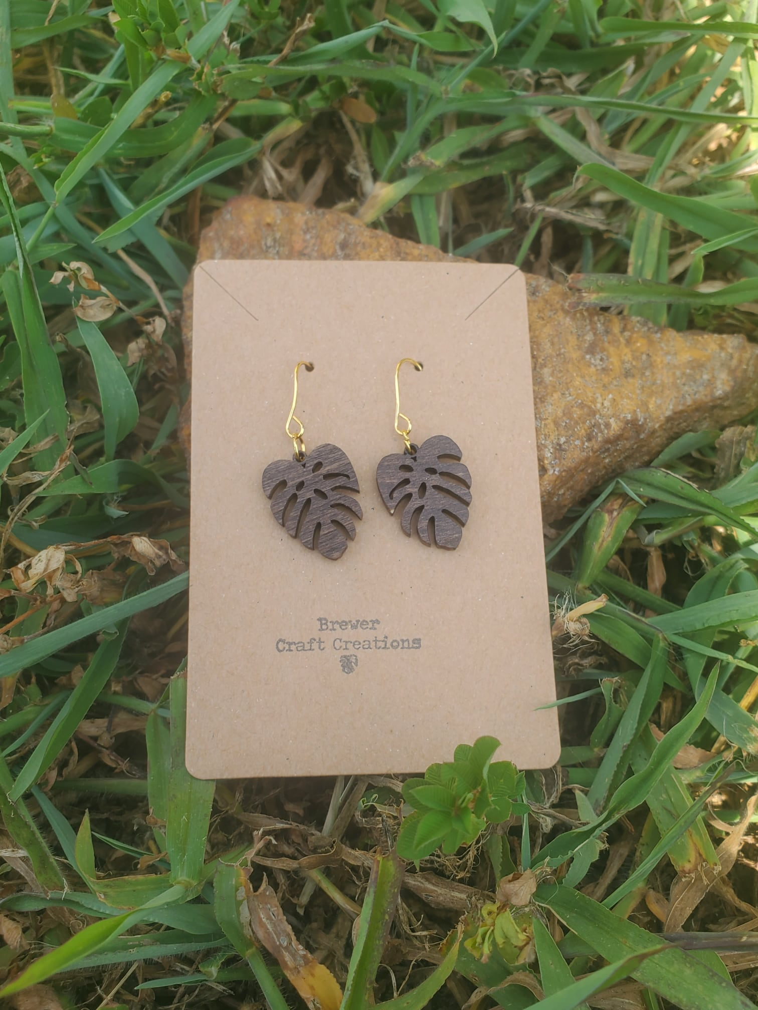 Signature monstera leaf earring on a cardboard backing being displayed on a rock in front of grass.